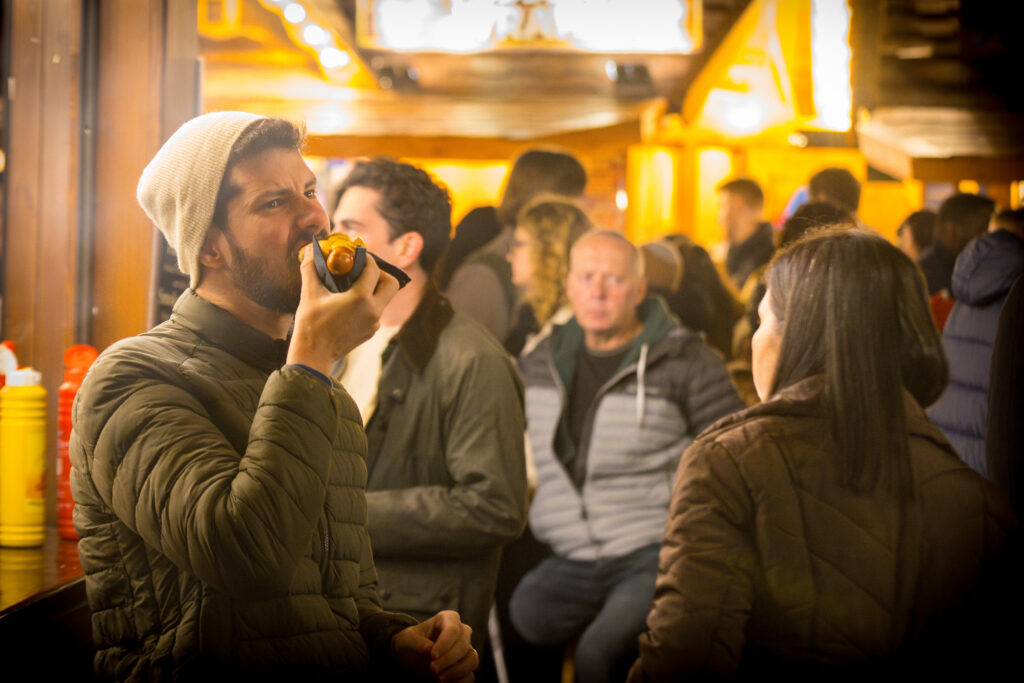 A man wearing a woolly hat eating a hot dog in a crowded street food market place.